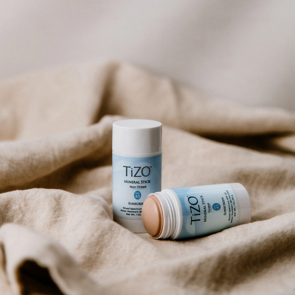 Two TiZO Mineral Stick tinted SPF45 sunscreen sticks on a beige fabric background, one upright and one lying down showing the tinted cream.